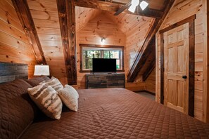 A wooden cabin bedroom with a bed, pillows, a TV, and a window. - Timberland Ridge | Hocking Hills, New, Hot Tub (Logan)