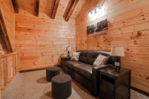L'intérieur d'un chalet en bois, avec un canapé en cuir, deux poufs, une table de chevet avec une lampe et un tableau encadré au mur.