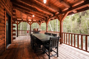 A covered wooden deck with a dining table and chairs, surrounded by a railing with a forest view.