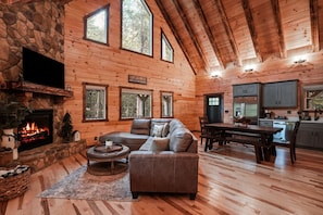 A log cabin interior with a stone fireplace, a sectional sofa, a dining table, and a kitchen area. - Timberland Ridge | Hocking Hills, New, Hot Tub (Logan)