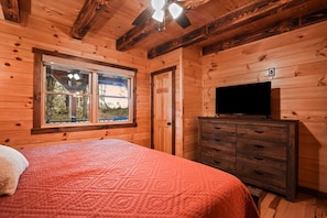 Un dormitorio de cabaña de madera con cama, cómoda, televisor y ventana con vistas.
