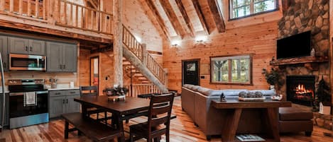 A wooden cabin interior with a dining area, kitchen, living room, and fireplace.