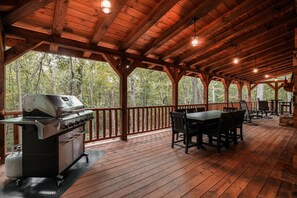 Une terrasse extérieure en bois, équipée d’un barbecue, d’une table à manger, de chaises et offrant une vue sur la forêt.