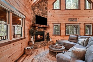 A cozy cabin interior with a stone fireplace, wooden walls, a leather sofa, and large windows.
