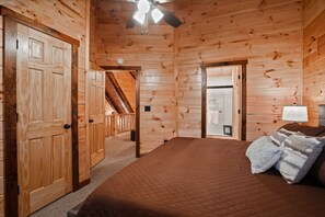 A wooden cabin interior with a bed, a ceiling fan, and a staircase leading to an upper level.