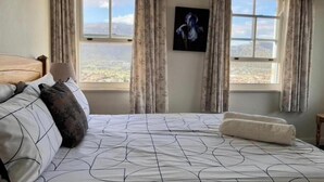 Une chambre à coucher avec un lit, des rideaux, une fenêtre donnant sur un beau paysage et un tableau encadré accroché au mur.