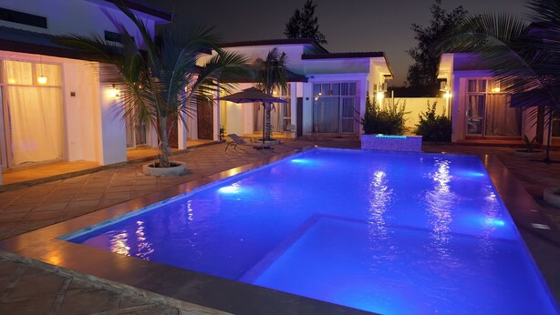 Una piscina iluminada con luces azules, rodeada de un patio con sillas y palmeras.