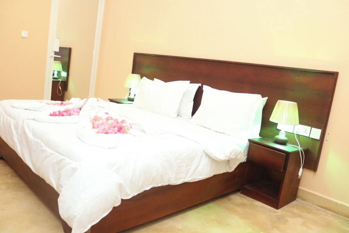 Una cama bien hecha con sábanas blancas y pétalos de flores rosa, un cabecero de madera, dos mesitas de noche con lámparas y un espejo en la pared.