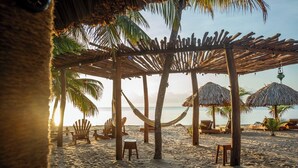 Una playa con sombrillas de paja, sillas de madera y hamacas bajo una pérgola de madera.