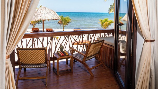 Una terraza de madera con dos sillas de mimbre, una mesa pequeña con fruta y un paraguas de paja.