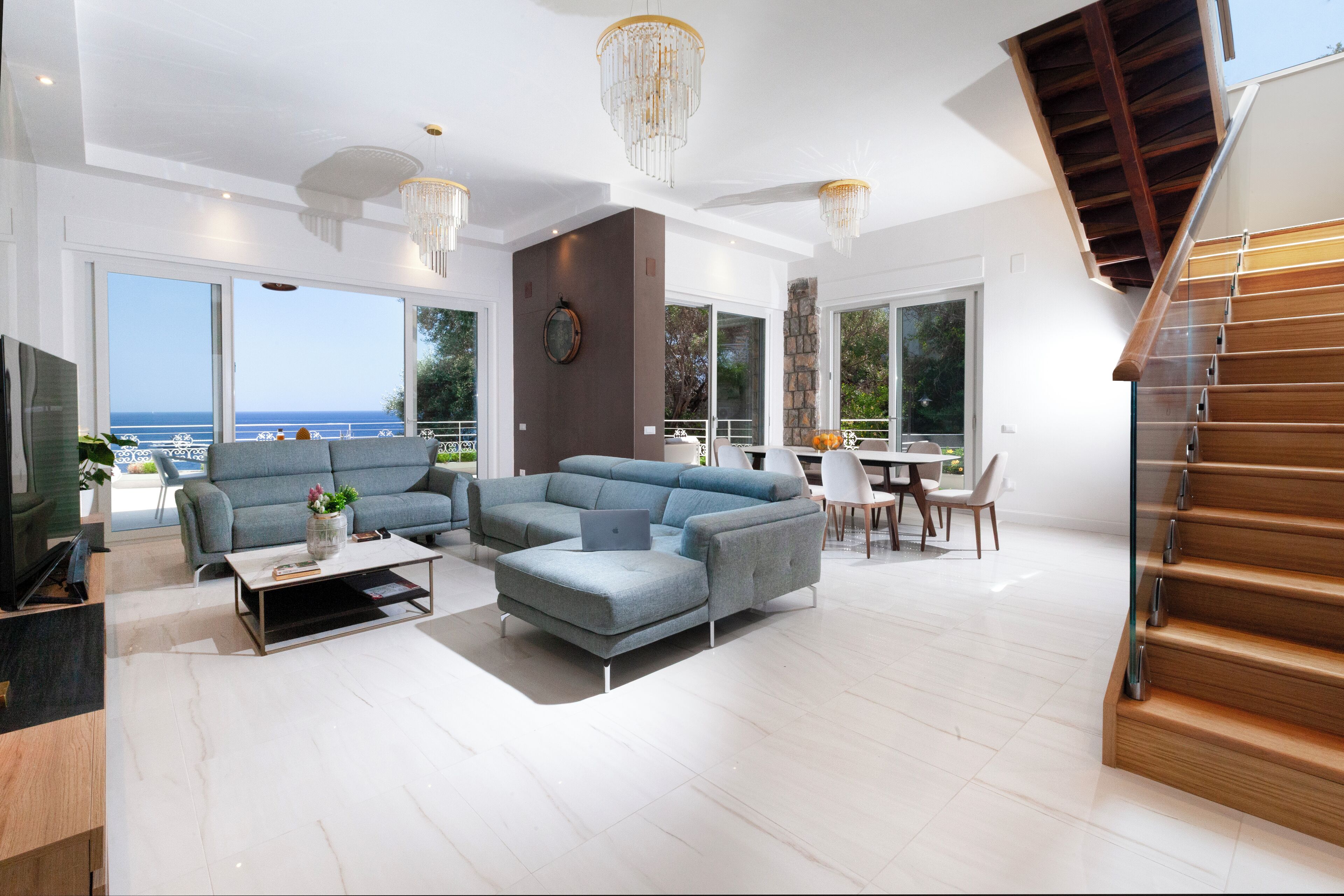 Een moderne woonkamer met een bank, salontafel en televisie. Een trap aan de rechterkant en een balkon met uitzicht op zee.