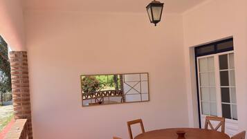 Eine Terrasse mit einem runden Holztisch und Stühlen, einem hängenden Laternenlicht, einem Fenster mit weißen Rahmen und einer Backsteinmauer.