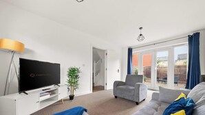 A living room with a flat-screen TV, a potted plant, a grey armchair, a sofa, and a large window with curtains. - Drakeway House, Reading (Berkshire)