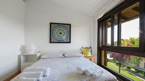 Ein Schlafzimmer mit einem Bett, einem Nachttisch, einer Lampe, einem gerahmten Kunstwerk und einem Fenster mit Blick auf Grünflächen.