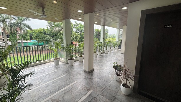 A covered outdoor area with potted plants, a tiled floor, and a view of a green space with trees. - Hotel Dhana Lakshmi Bliss (Dewas)