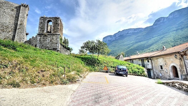 A paved road leading to a historic stone building with a tower, surrounded by greenery and mountains.