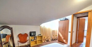 Un dormitorio con cama, un armario de madera, un televisor sobre una estantería, una silla y una ventana con cortinas.