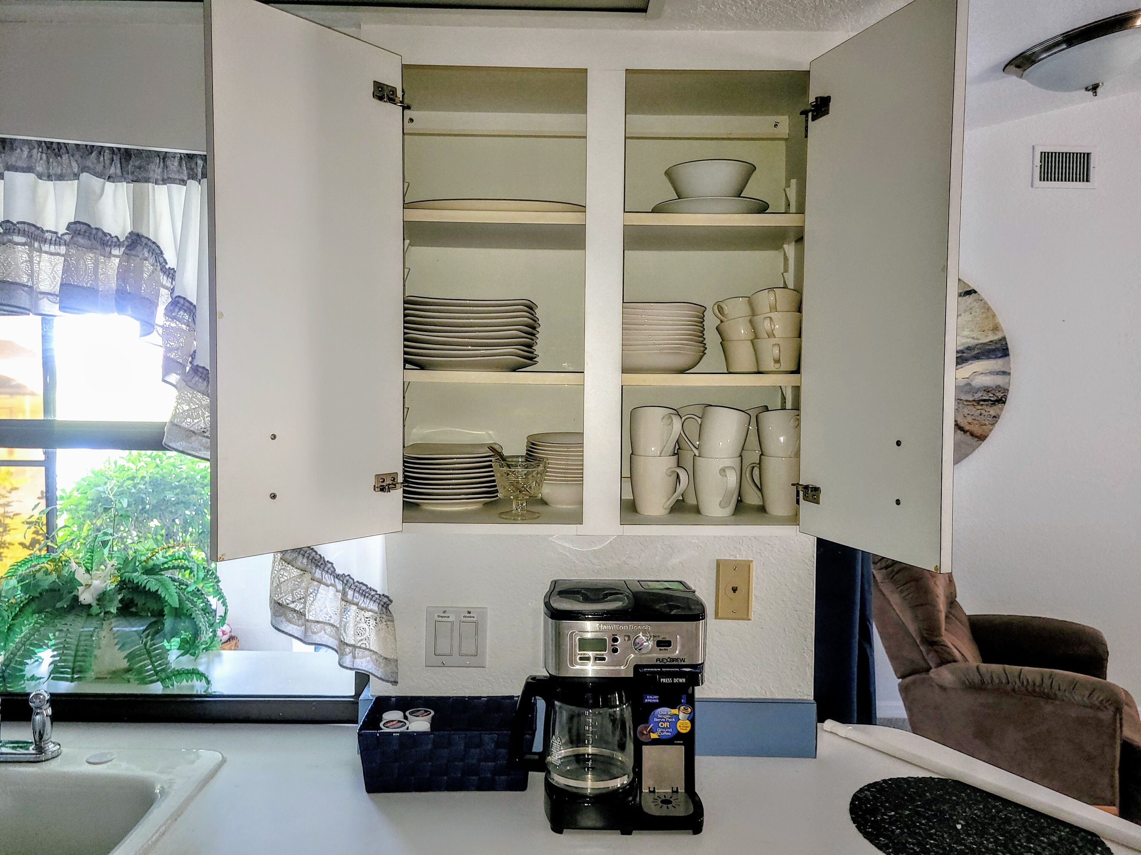 A kitchen with a cabinet containing plates and cups, a coffee maker, and a window with curtains.
