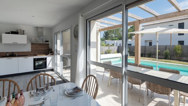 Une cuisine moderne avec des meubles blancs, un coin repas comprenant une table et des chaises, et une vue sur une piscine à travers de grandes fenêtres.
