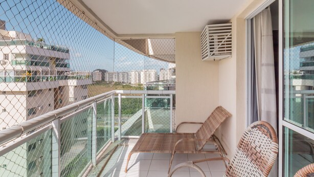 Een balkon met rieten stoelen, een glazen hekwerk en uitzicht op gebouwen.