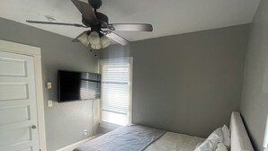 A bedroom with a bed, a ceiling fan, a TV mounted on the wall, and a window with blinds.