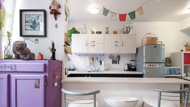 Une cuisine avec des meubles blancs, un réfrigérateur violet et un petit coin repas avec deux chaises.