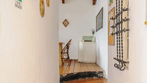 Un estrecho pasillo con suelo de baldosas que lleva a una habitación con una puerta blanca. Hay cuadros enmarcados en la pared, una señal de salida verde y un estante decorativo de metal.