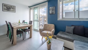 Ein modernes Wohnzimmer mit Essbereich, einer Couch und einem Sessel. Ein großes Fenster bietet Blick auf Gebäude und einen Balkon.