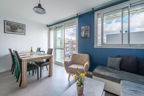 Una sala moderna con comedor, un sofá y una silla. Hay un ventanal con vistas a edificios y un balcón.