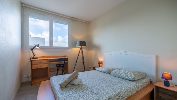 Ein Schlafzimmer mit Bett, Schreibtisch, Stuhl, Lampe und Fenster.