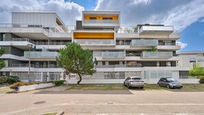 A modern multi-story building with balconies, a central yellow section, and a parking area with cars.