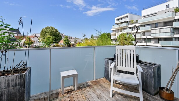 A balcony with a white chair and a small table, overlooking a modern building and a tree.
