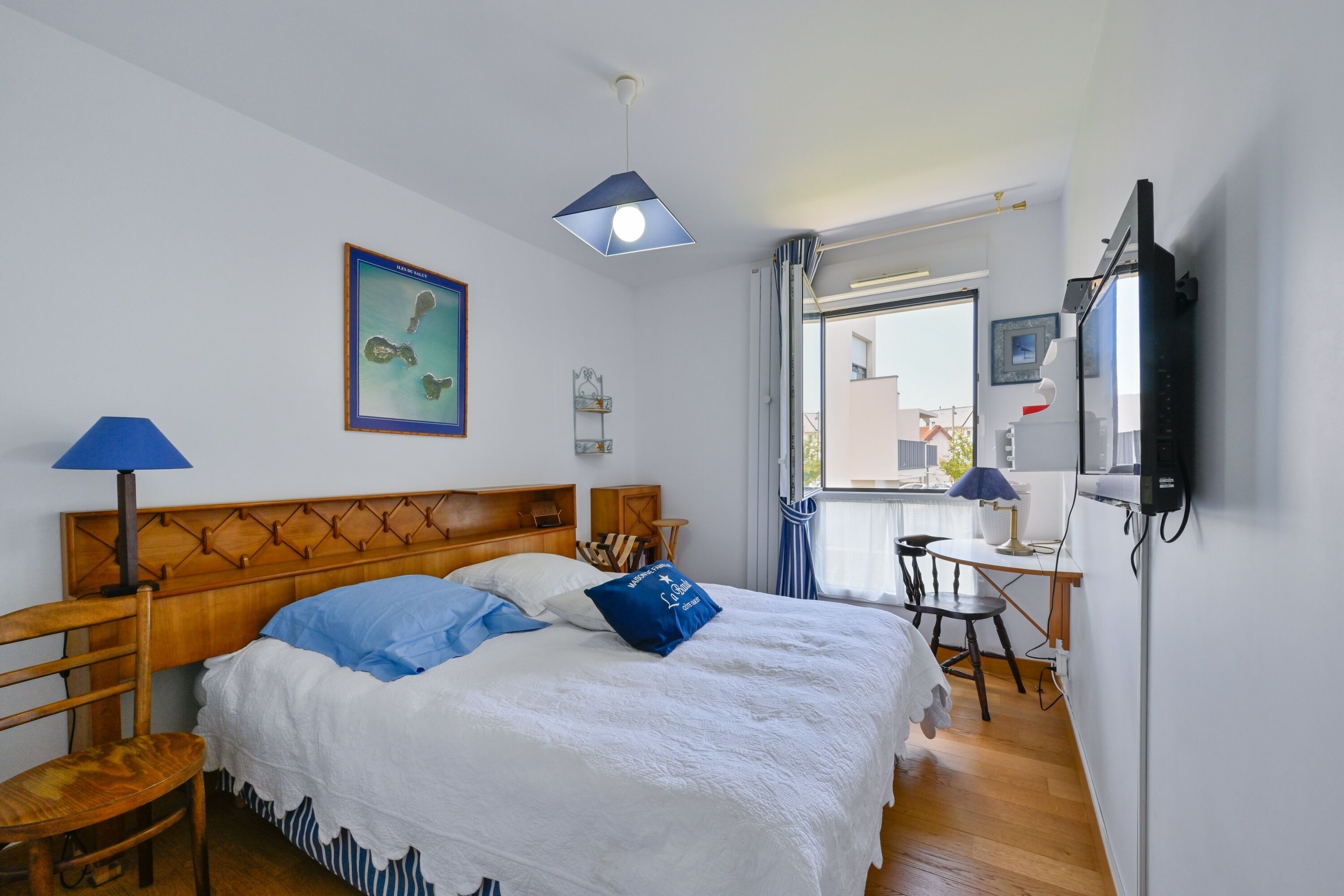 A hotel room with a wooden headboard, a bed with white and blue bedding, a desk with a chair, a television, and a window with curtains.