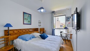 A hotel room with a wooden headboard, a bed with white and blue bedding, a desk with a chair, a television, and a window with curtains.