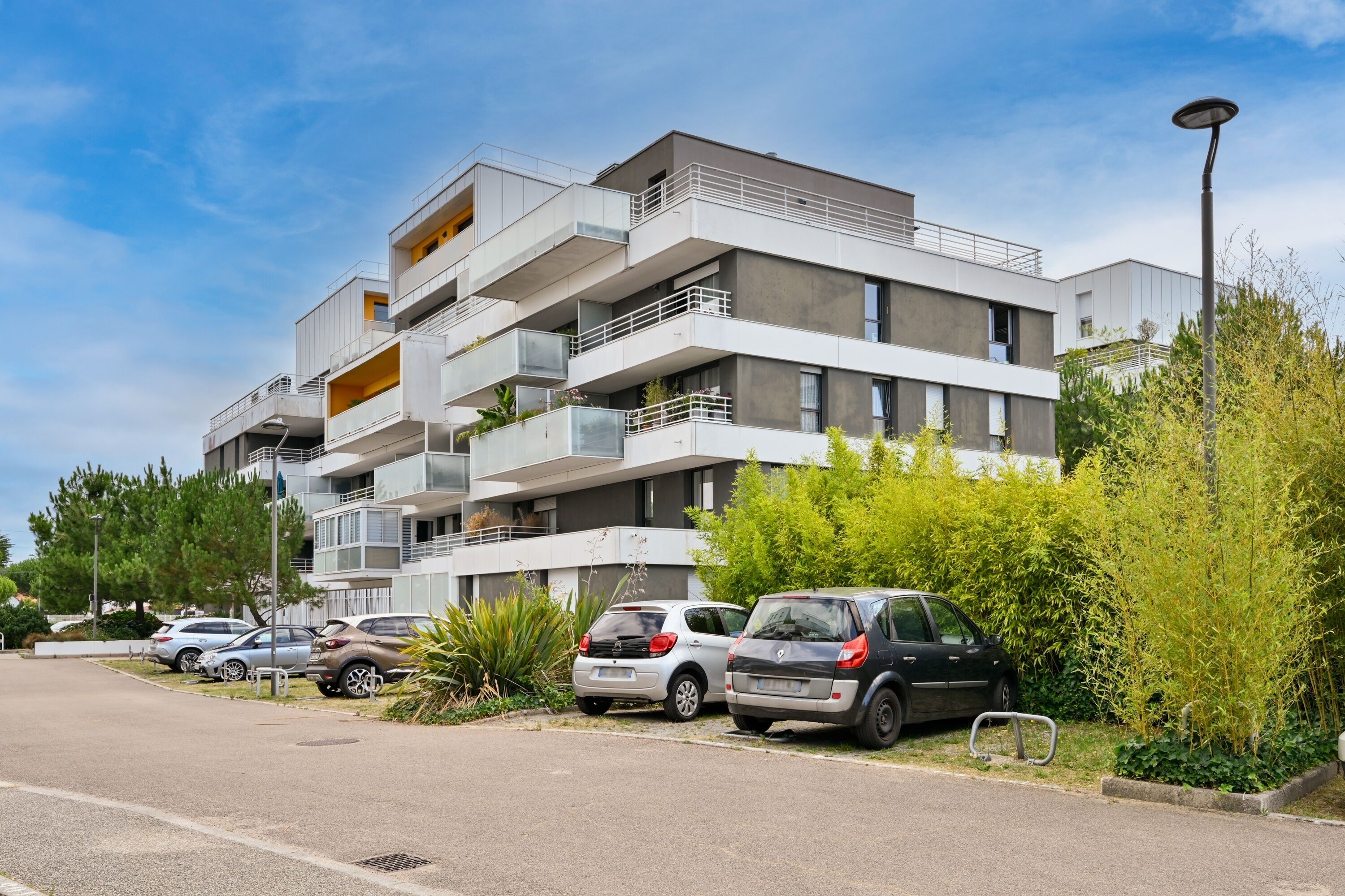 A multi-story residential building with balconies, a parking area with several cars, and surrounding greenery.