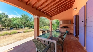 A covered patio with a glass-top table and chairs, a barbecue grill, and a view of the surrounding landscape.