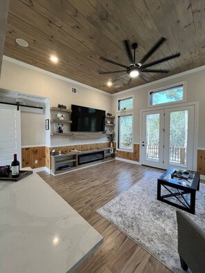 A spacious living area with a ceiling fan, a flat-screen TV, wooden flooring, a fireplace, and large windows.