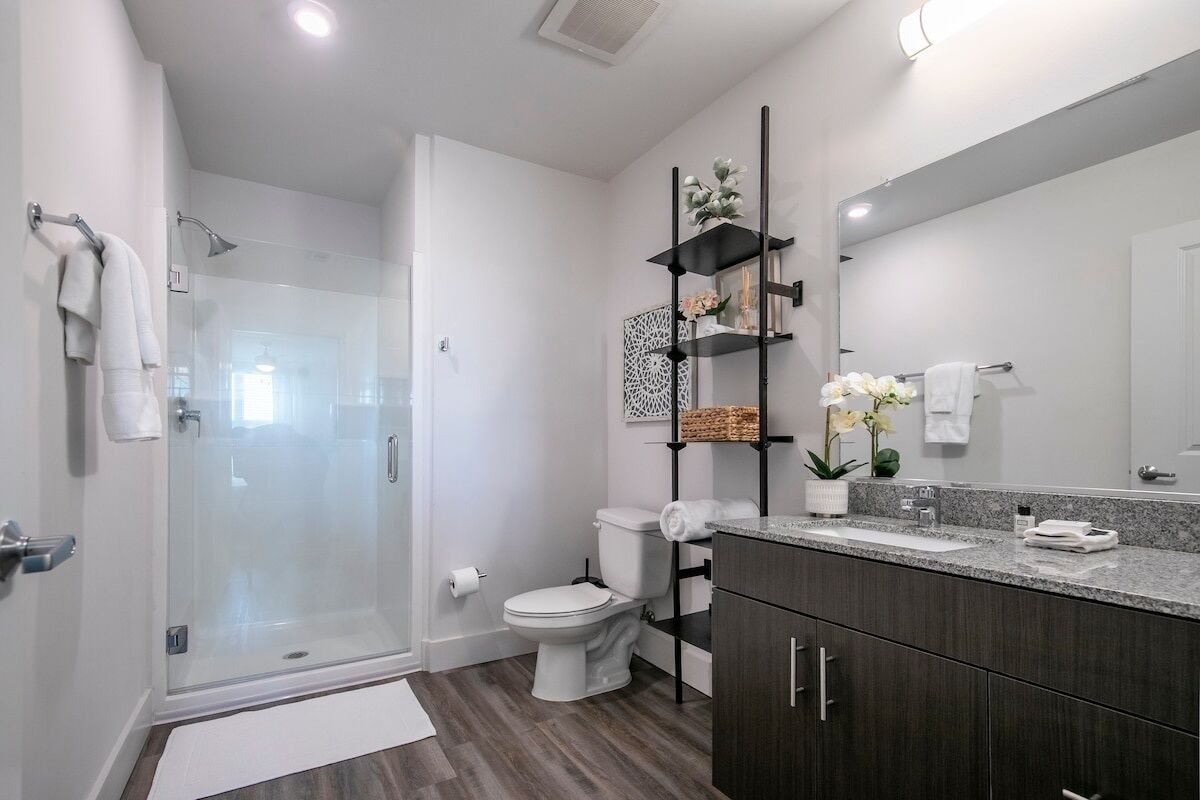 A modern bathroom with a glass shower enclosure, a toilet, a vanity with a large mirror, and a shelf with decorative items.