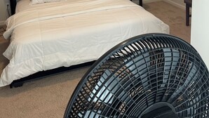 Une chambre à coucher avec un lit, une tête de lit noire recouverte de tissu, du linge de lit blanc et un ventilateur sur pied.