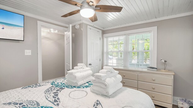 Une chambre à coucher avec un lit, un ventilateur de plafond, une commode, une fenêtre avec des stores et un téléviseur fixé au mur.