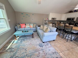 A living room with a sofa, coffee table, and a rug. There is a kitchen area with cabinets and a microwave.