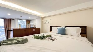A hotel room with a bed, a bedside table, a hanging clothes rack, a window with curtains, and a view of the city. - Quiet And Strategic Studio Loft At Apple 1 Condovilla Apartment (Jakarta)