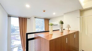 A modern kitchen with wooden cabinets, a white vase with greenery, a lamp, and a window with curtains. - Quiet And Strategic Studio Loft At Apple 1 Condovilla Apartment (Jakarta)