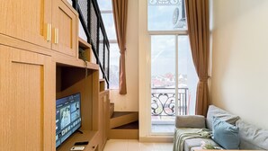 A living room with a TV on a wooden cabinet, a sofa with cushions, and a balcony with a railing. - Quiet And Strategic Studio Loft At Apple 1 Condovilla Apartment (Jakarta)