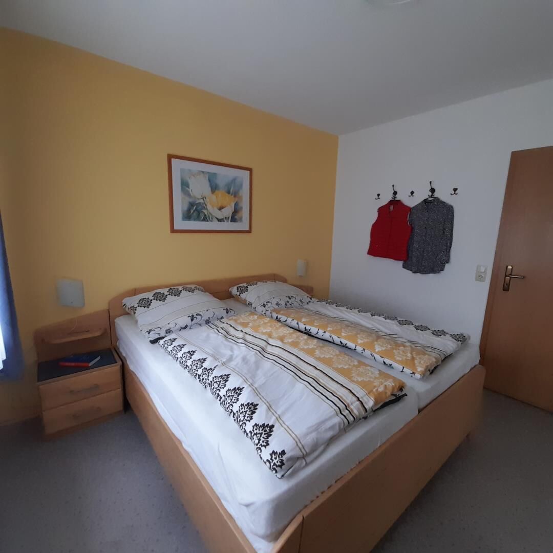 Un dormitorio con cama, mesita de noche, perchero en la pared y una puerta.