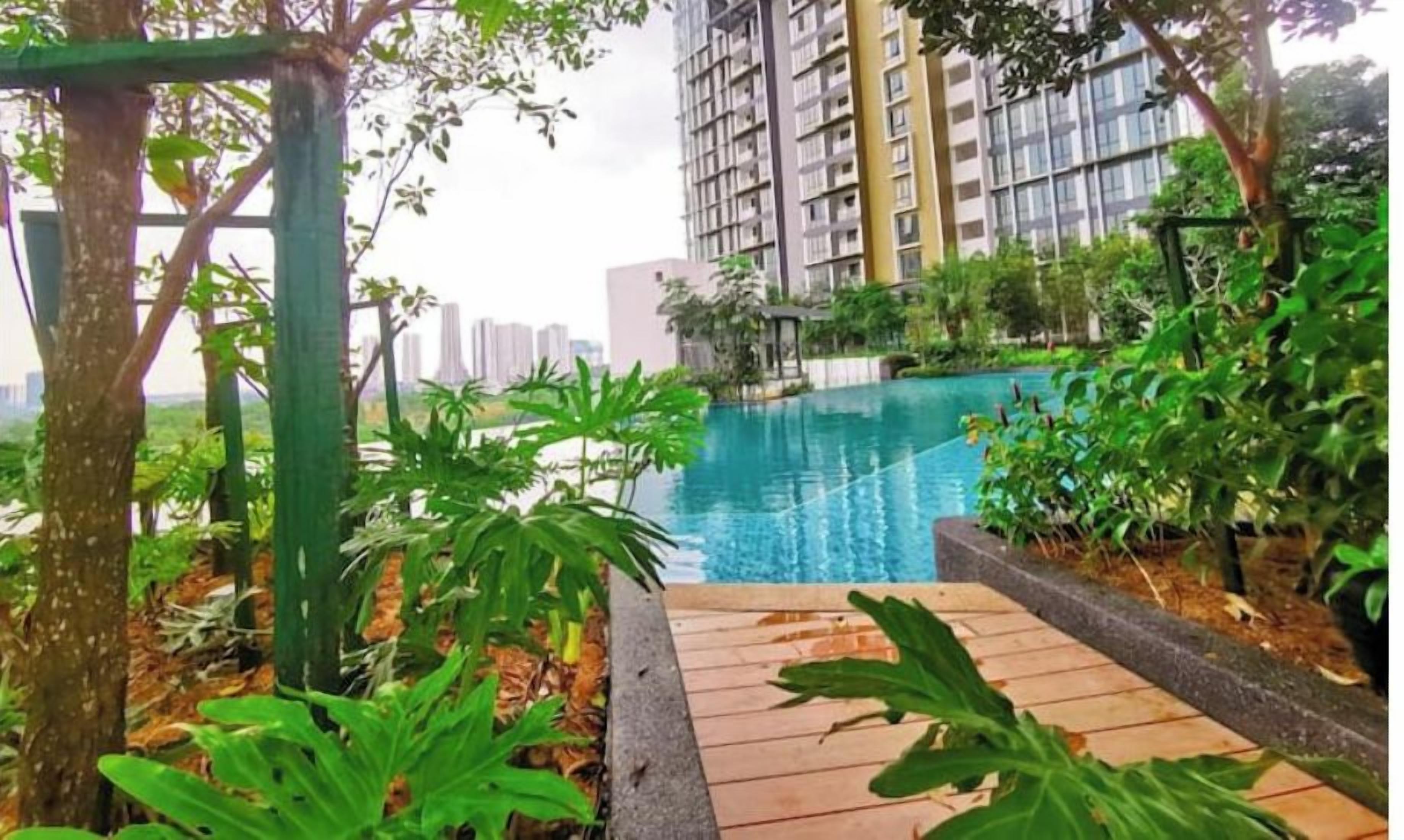 A pool area with a wooden deck, surrounded by lush greenery and tall buildings in the background.