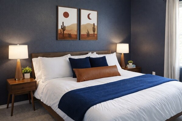A neatly made bed with white and blue bedding, two bedside lamps, and two framed desert-themed wall art pieces.