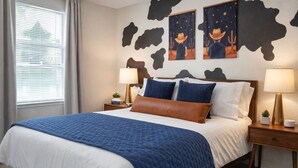 Quarto de hotel com uma cama grande, duas mesinhas de cabeceira, um criado-mudo, uma janela com persianas e obras de arte na parede retratando cowboys e cactos.
