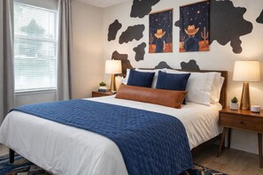Una habitación de hotel con una cama grande, dos mesitas de noche, un tablero de noche, una ventana con persianas y arte en la pared que representa vaqueros y cactus.