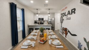 Uma área de jantar com uma longa mesa de madeira posta para o café da manhã, uma cozinha ao fundo e um mural na parede com um cacto e as palavras 'TOLAR' e 'Texas'.
